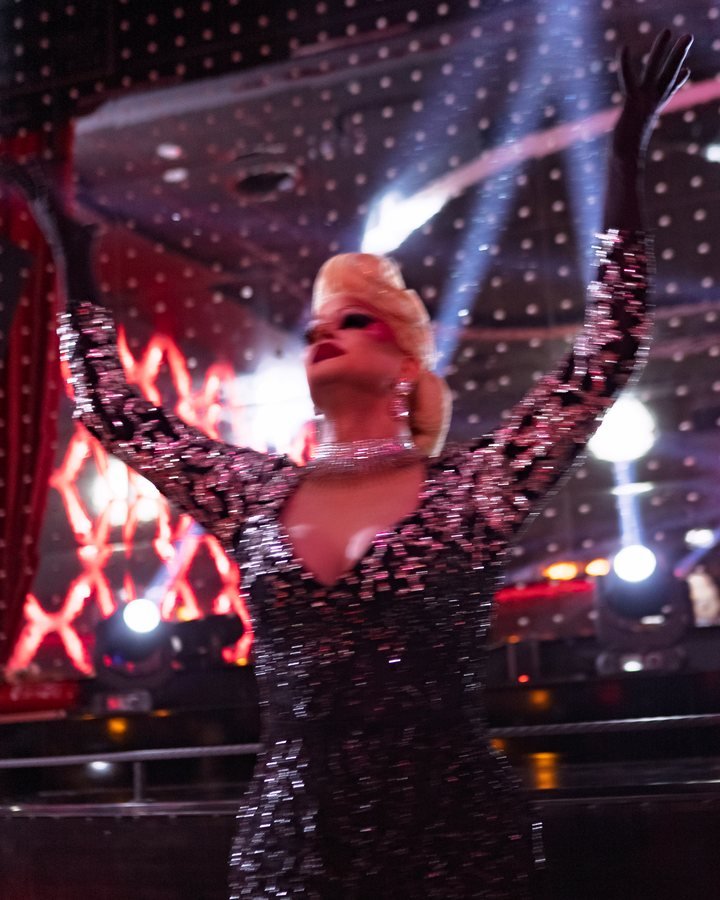 A cabaret singer in a sequinned gown, arms raised, on the Stable 54 stage.