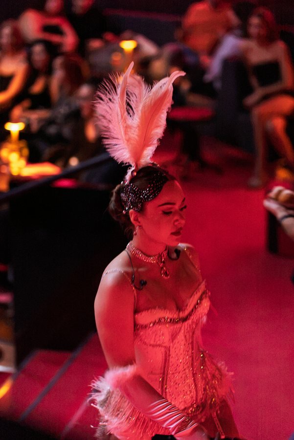 A showgirl in a feather headdress lit in deep red among the guests.