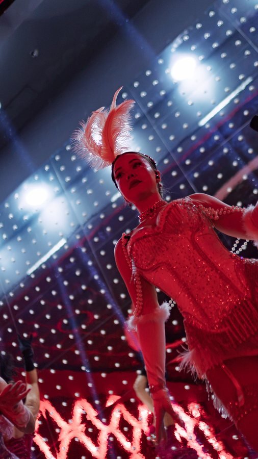 A dancer in a red fringed costume and feathers under the stage lights.