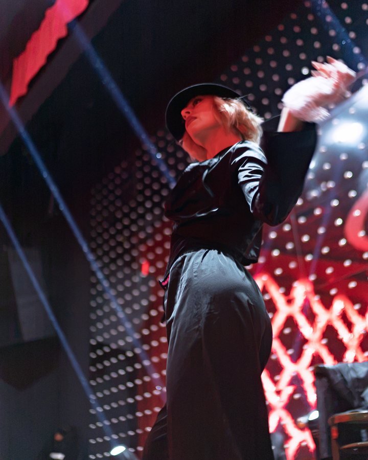 A performer in a black hat and coat, one arm raised, under a stage light.