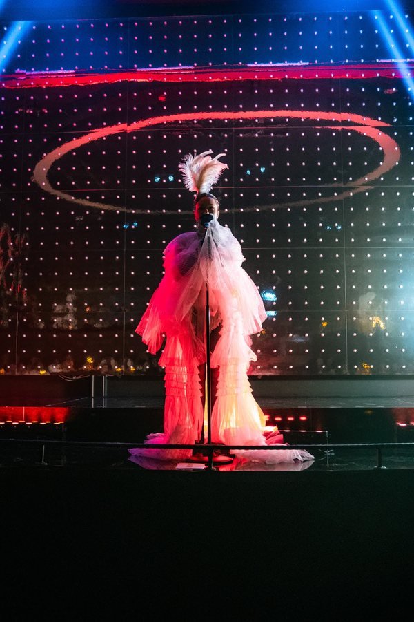 A singer in a white tulle gown and feather headdress at the microphone on the Stable 54 stage.