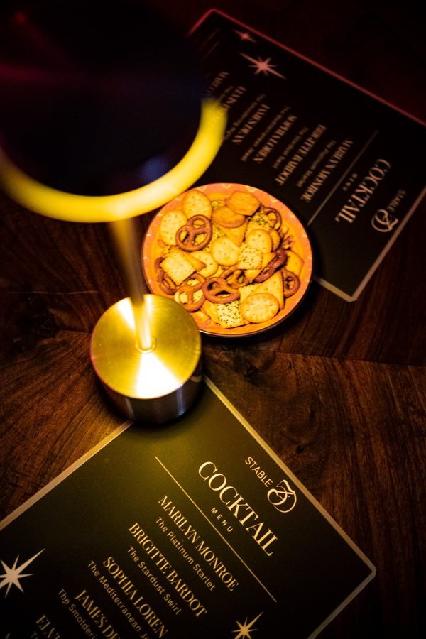 The Stable 54 cocktail menu beside a brass table lamp and a bowl of snacks.