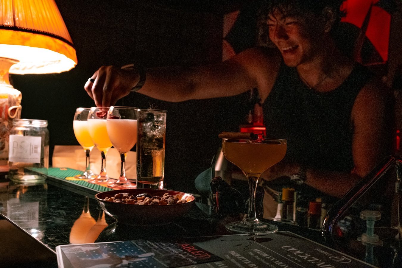 A smiling bartender garnishing a row of cocktails under a lamp.