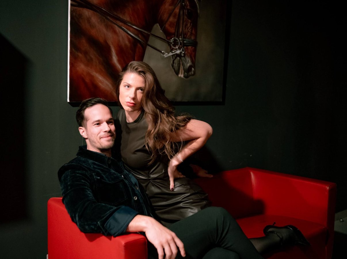 Two guests on a red sofa beneath a painting of a horse.