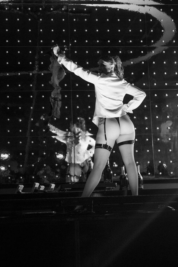 A burlesque performer in a white shirt and stockings posing on stage, in black and white.