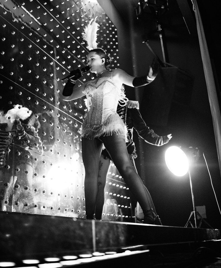 A showgirl in a corset and feathers singing on stage, in black and white.