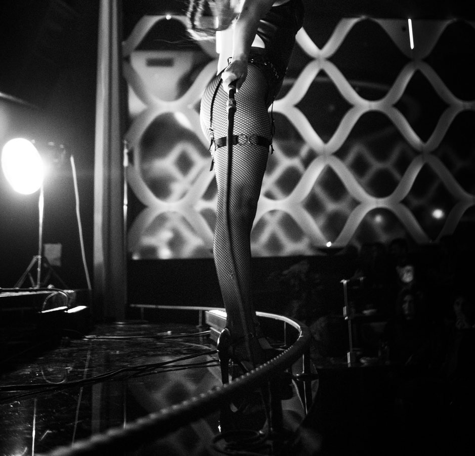 A burlesque performer in fishnets on the edge of the stage beside the lattice wall, in black and white.